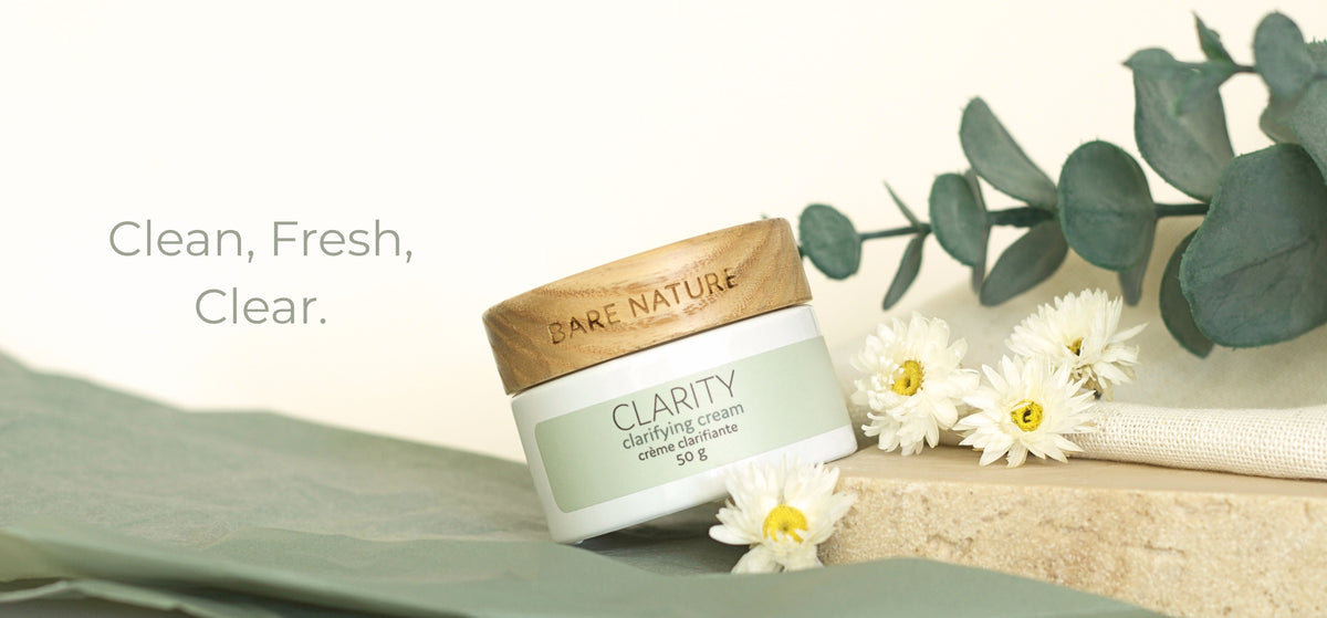 A jar of Bare Nature Clarity Cream is styled on a light beige surface with white daisies, green eucalyptus leaves, and a soft green fabric. The text 'Clean, Fresh, Clear.