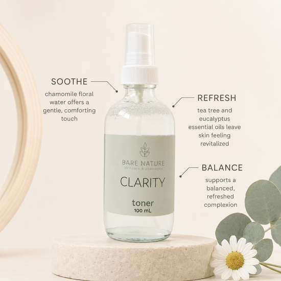 A clear glass spray bottle of Bare Nature Clarity Toner with a white pump and pale green label, displayed on a stone pedestal with eucalyptus leaves and Chamomile blossom.