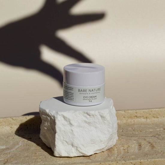 A small white cosmetic jar labeled 'BARE NATURE Eye Cream' sits on a rough white stone pedestal against a warm beige wall, with a hand shadow cast across the background.