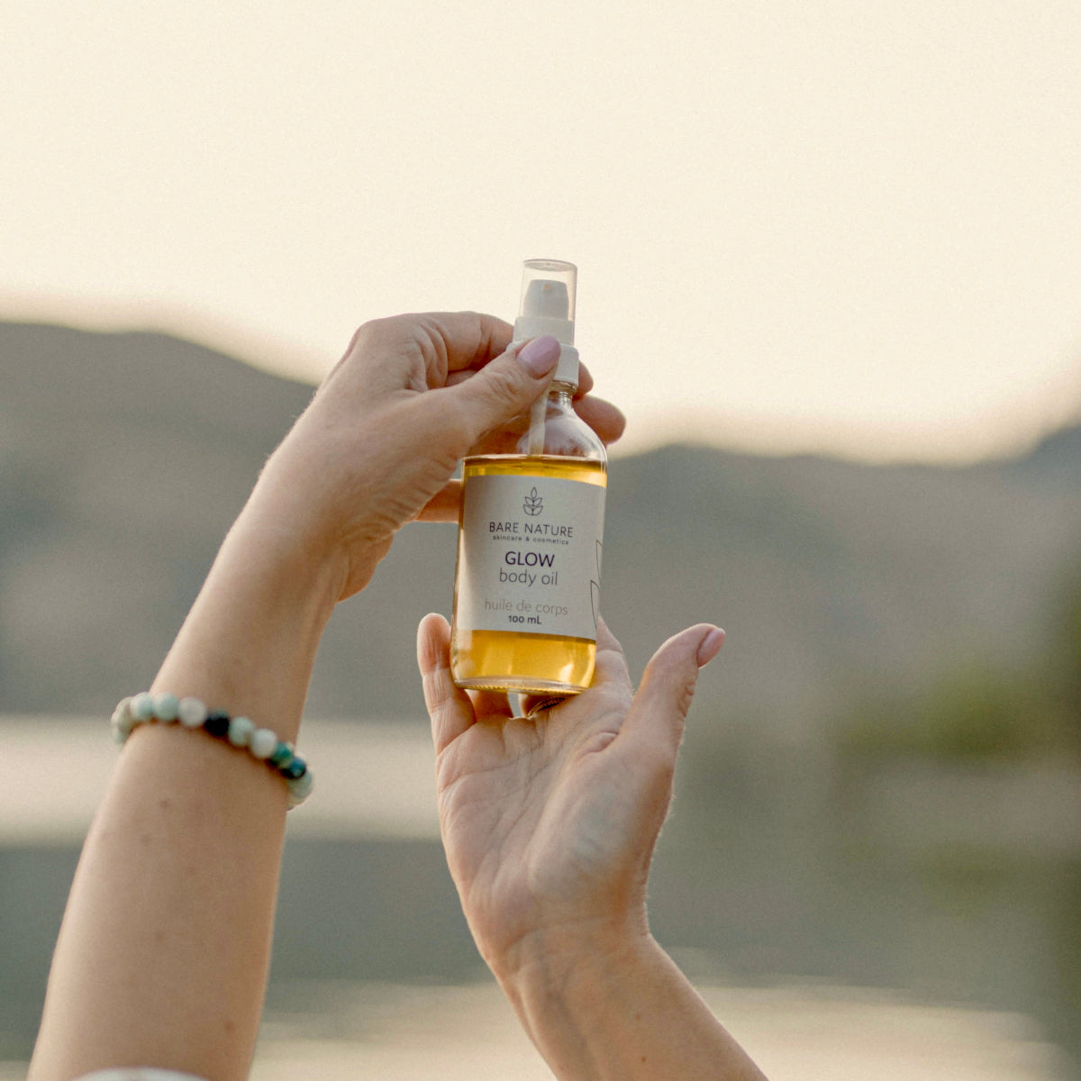 Person holding a bottle of 'Glow' body oil with a scenic background
