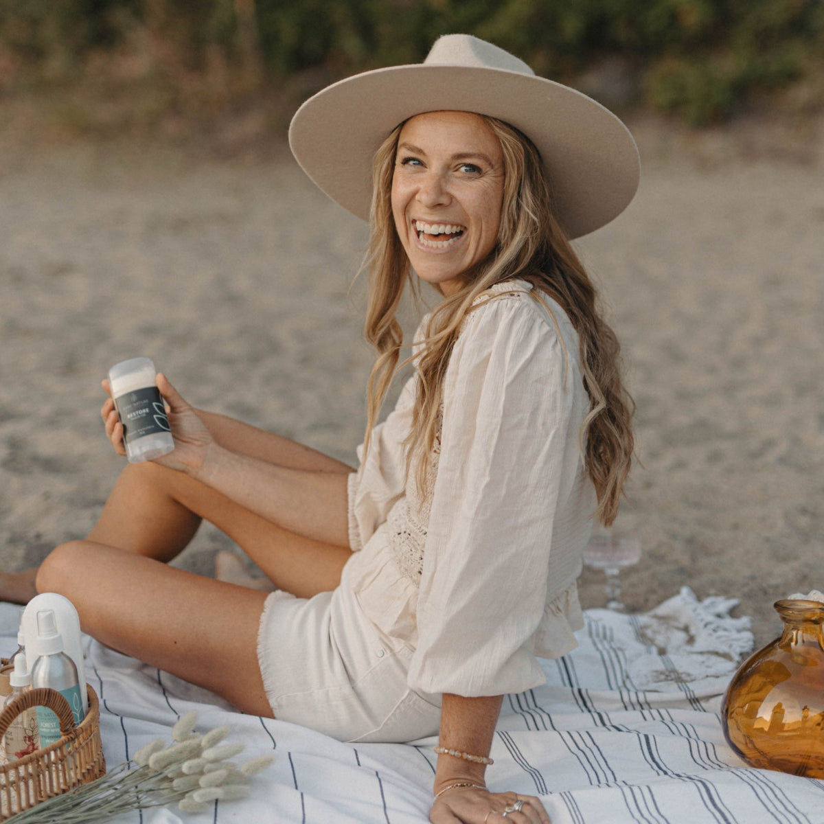 A woman sitting on a picnic blanket outdoors, smiling and holding a Bare Nature Restore muscle rub stick. She is wearing a wide-brimmed beige hat, a cream blouse, and shorts, with a basket, dried flowers, and an amber glass bottle nearby on a sandy beach-like setting.