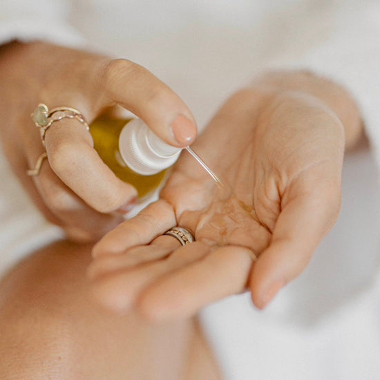 A close-up of a person's hands dispensing Bare Nature Glow Body Oil from a small pump bottle into their palm, with rings visible on their fingers.