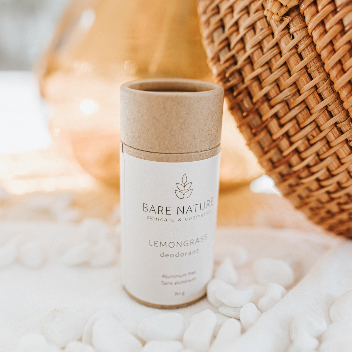 A cylindrical tube of Bare Nature Lemongrass Deodorant standing upright on white pebbles, with a kraft paper lid and a white label. A warm, softly blurred background includes a woven basket and a glass vessel.