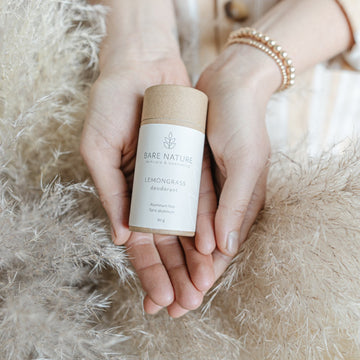 A person's hands holding a cylindrical cardboard container of Bare Nature Lemongrass Deodorant, set against a soft beige fluffy background.