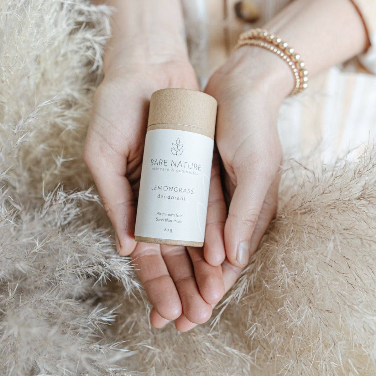 A person's hands holding a cylindrical cardboard container of Bare Nature Lemongrass Deodorant, set against a soft beige fluffy background.