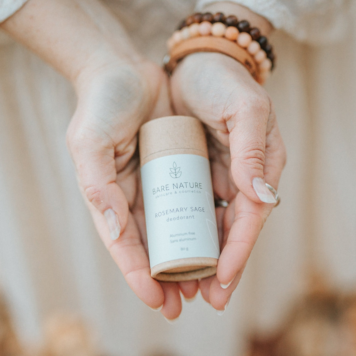 A person holds a Bare Nature Rosemary Sage deodorant stick in both hands. The product is in a natural kraft paper tube with a white label.