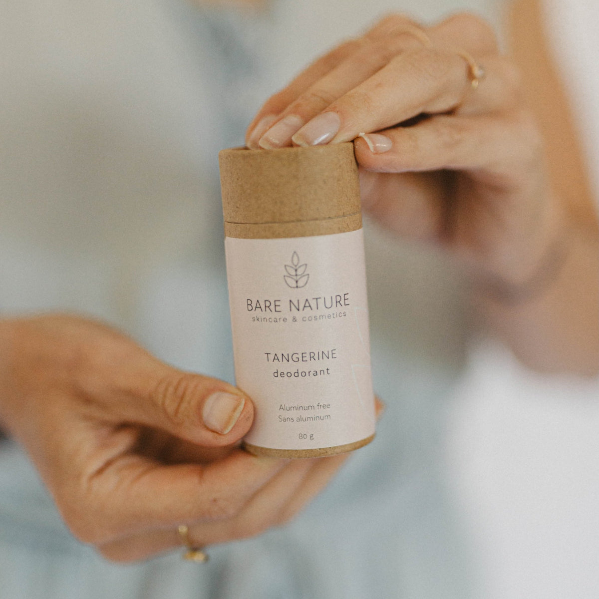 A person holds a small cylindrical deodorant container with a kraft paper top and a white label reading BARE NATURE TANGERINE deodorant. The background is softly blurred and light-colored.