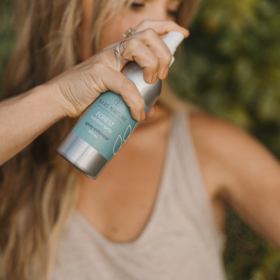 A woman outdoors holds a silver spray bottle labeled Bare Nature Forest  Spray, spraying it while wearing a beige tank top against a blurred green background.