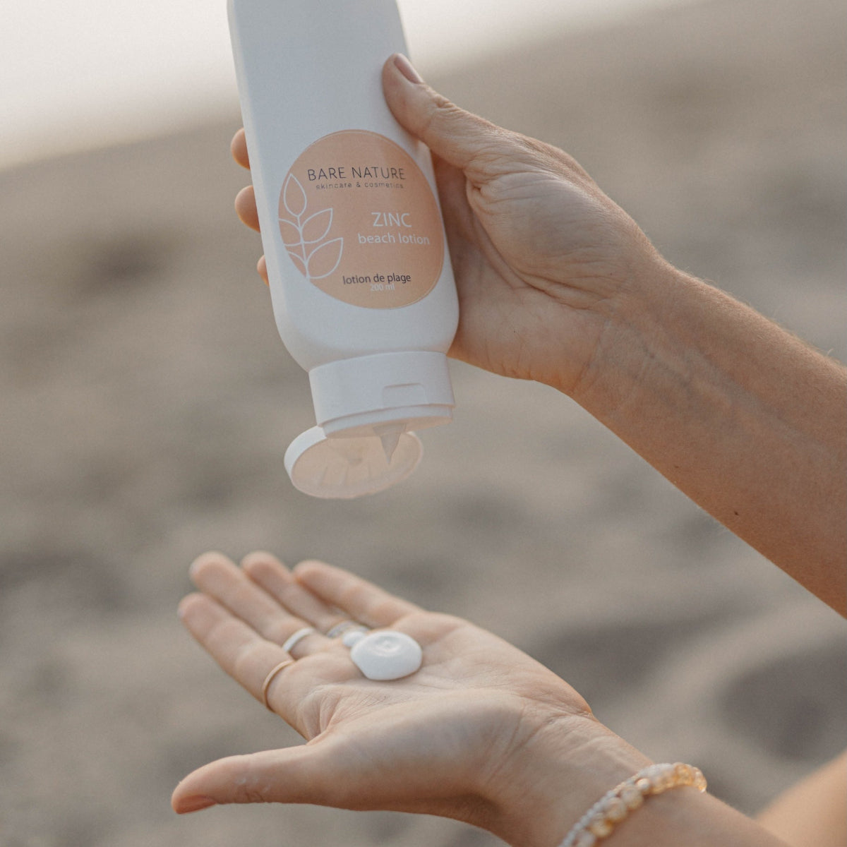 A person at the beach applies lotion from a white bottle labeled BASE NATURE ZINC. The bottle is held in one hand while the other hand receives a small dollop of the white cream.
