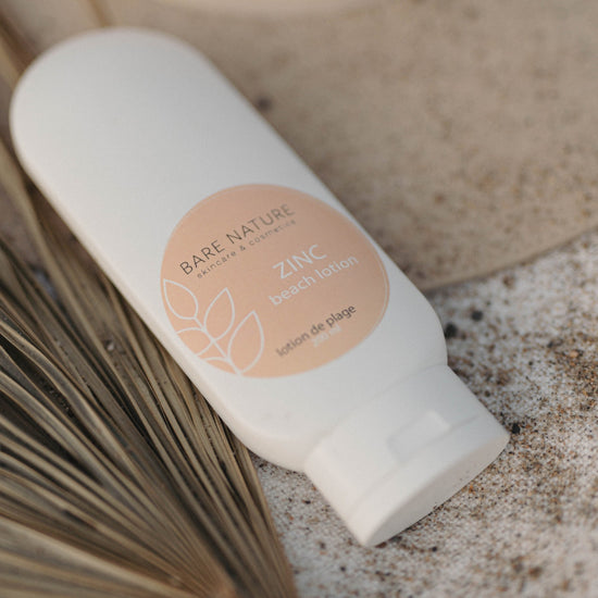 A white bottle of Bare Nature Zinc Beach Lotion resting on a textured stone surface with dried palm leaves, showing the peach-colored circular label.