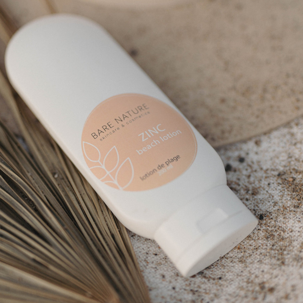 A white bottle of Bare Nature Zinc Beach Lotion resting on a textured stone surface with dried palm leaves, showing the peach-colored circular label.
